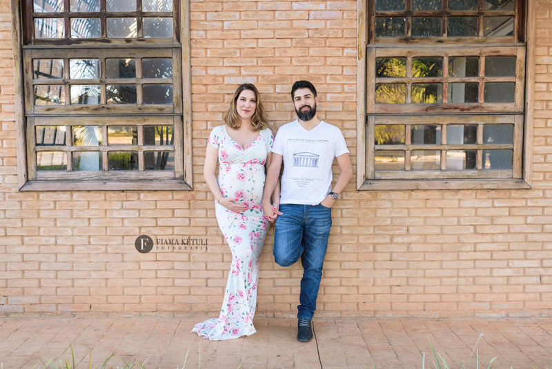Casal fazendo ensaio de gestante em parede de tijolinhos. Gestante com vestido longo florido importado
